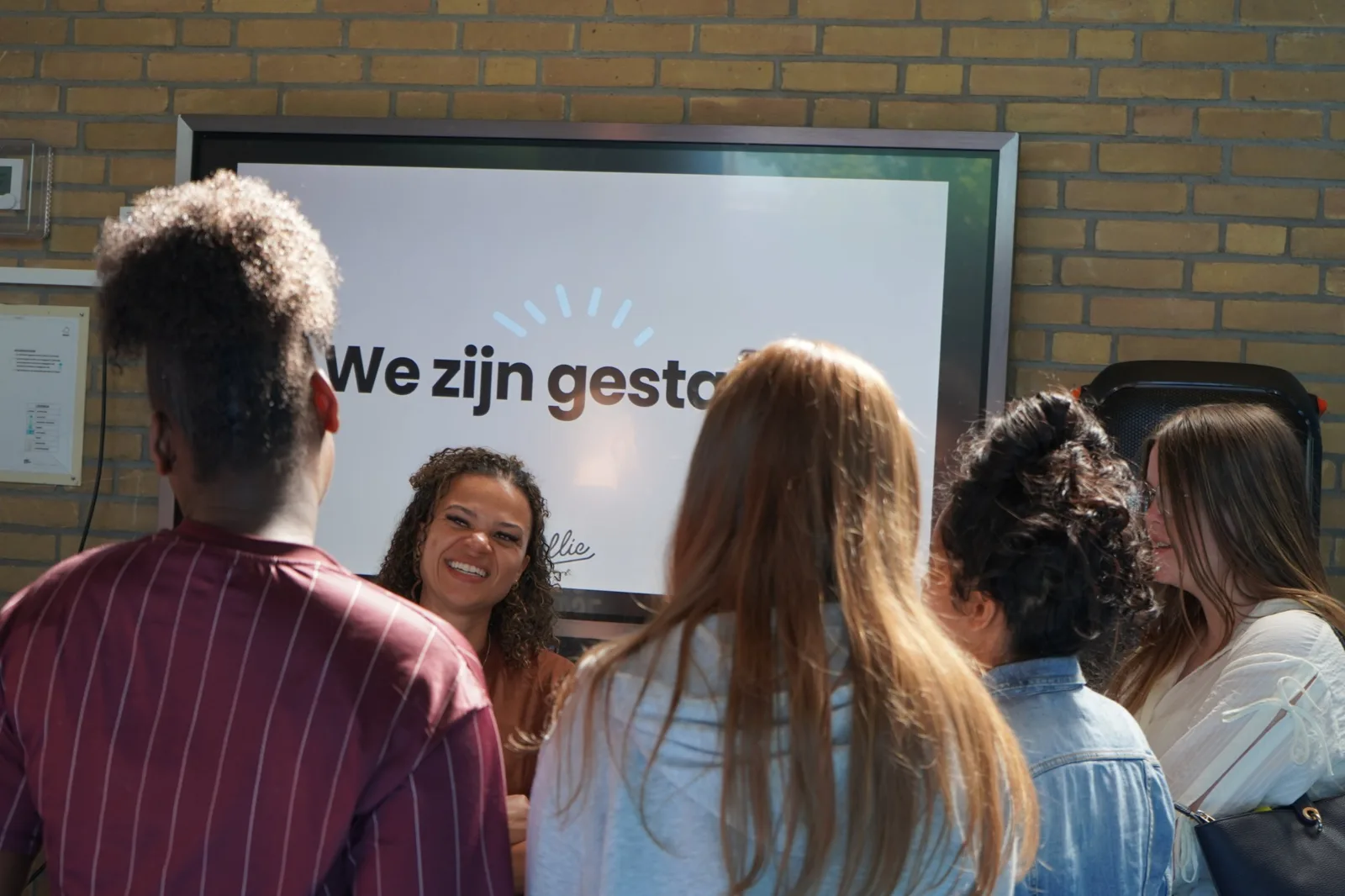 Trainer van Ollie lacht met een groep studenten voor een scherm met We zijn gestart
